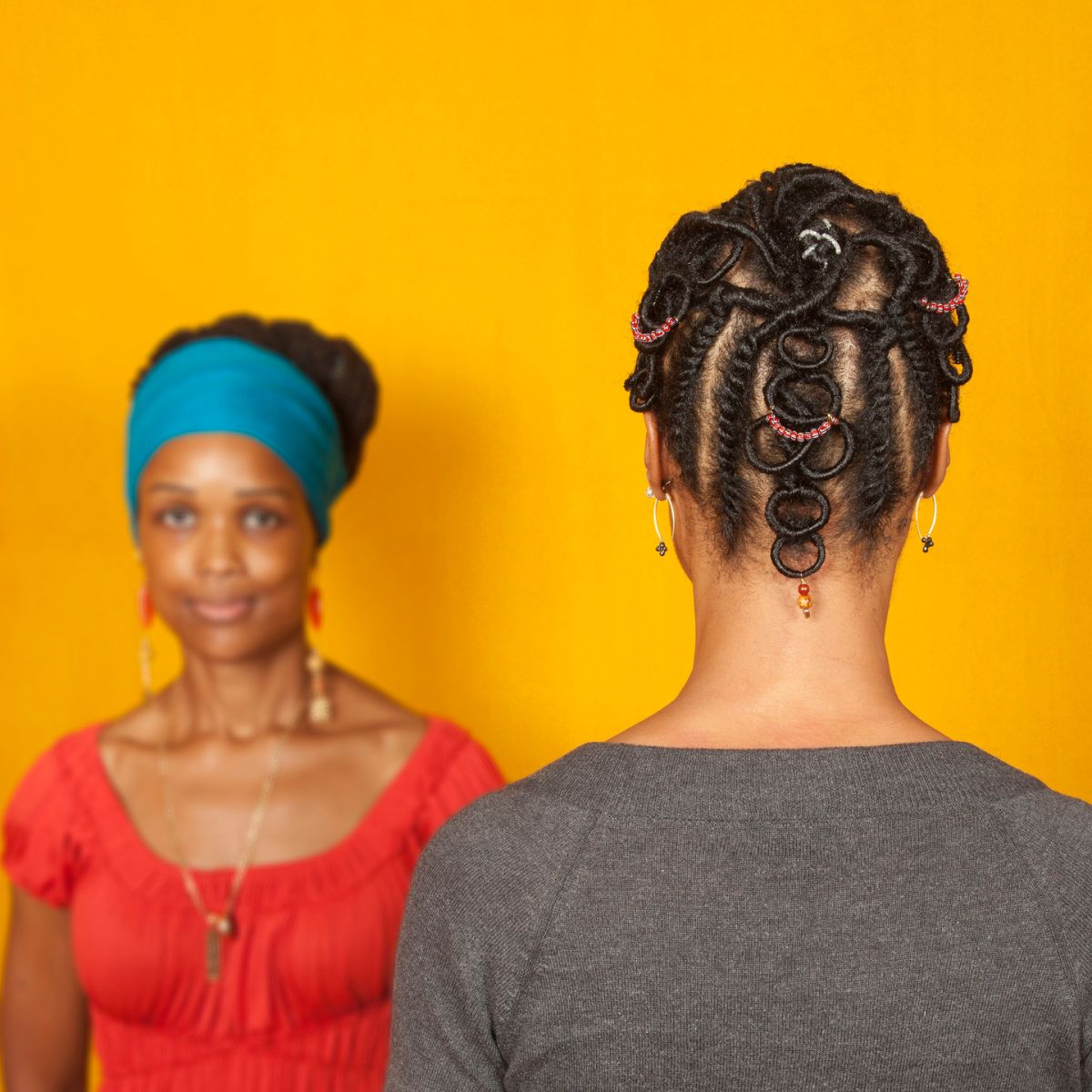 Two medium-dark skinned adult women stand in front of a goldenrod background. The woman on the left faces the viewer and wears a teal head wrap and a red top. The woman on the right faces away from the viewer, showcasing her dark hair braided into an elaborate pattern with colorful beads.