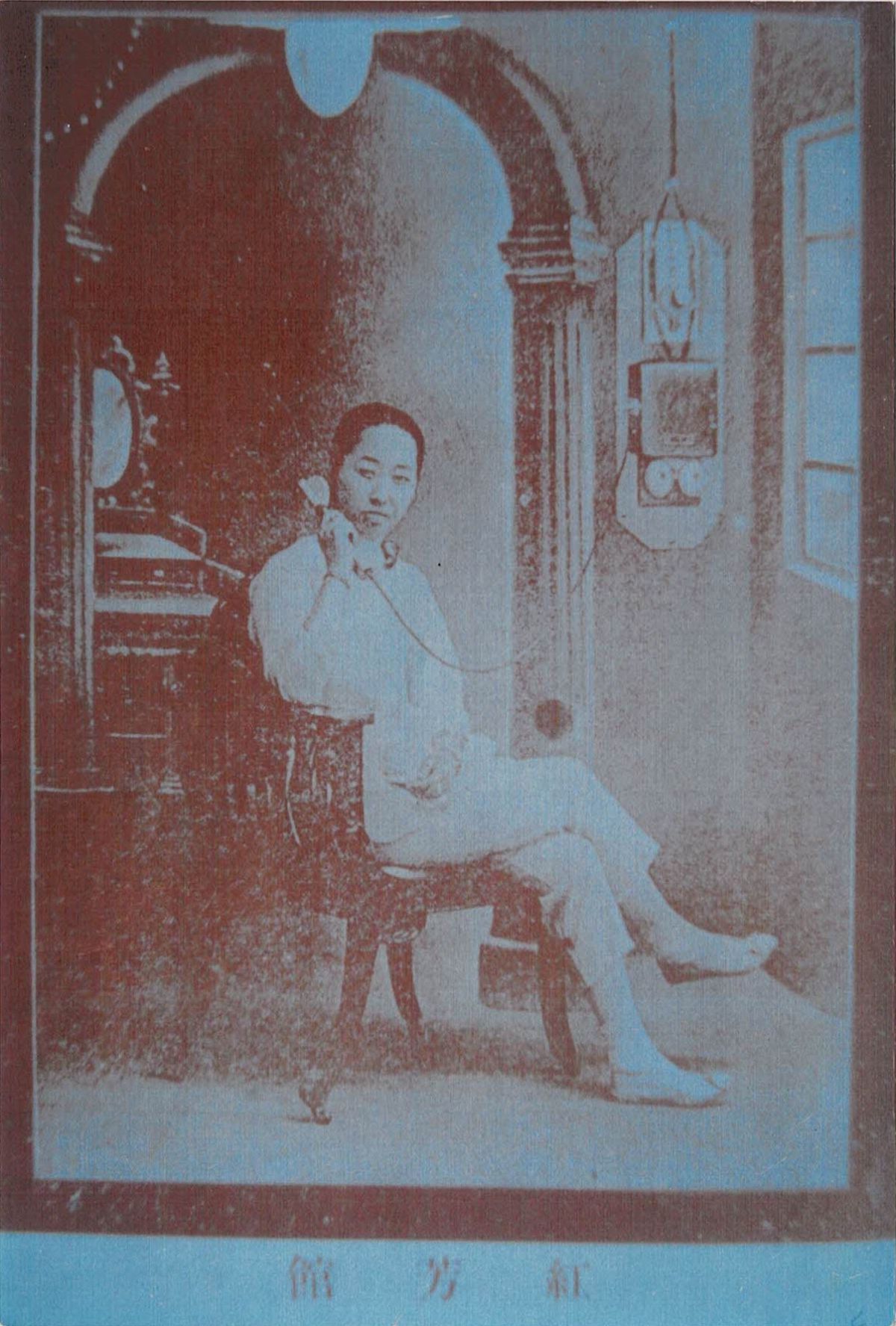 In a black-and-white image, an Asian woman poses while wearing a white blouse, pants, and shoes. She holds the receiver of a wall phone to her right ear, crosses one leg over the other, and smiles slightly at the camera.