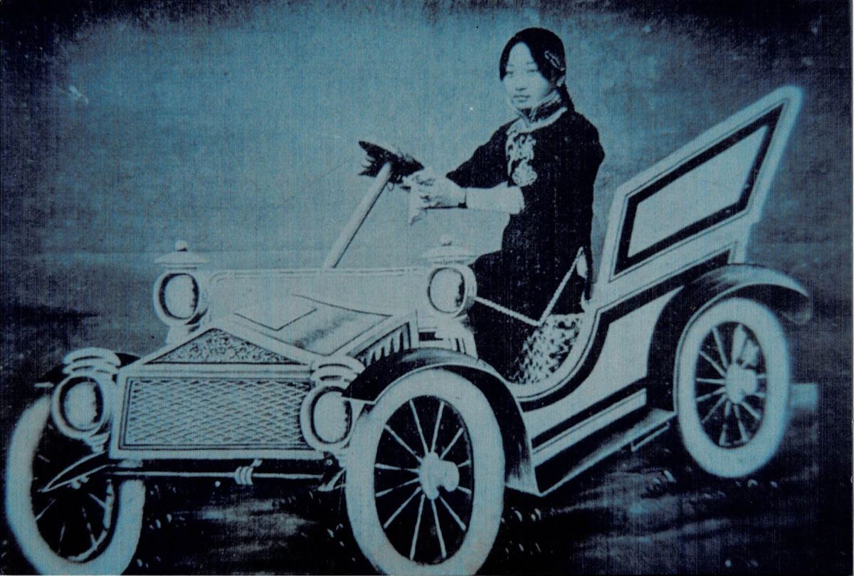 A black-and-white image shows a young Asian woman in black attire sitting in an early twentieth-century white prop car. Her hands are on the steering wheel.