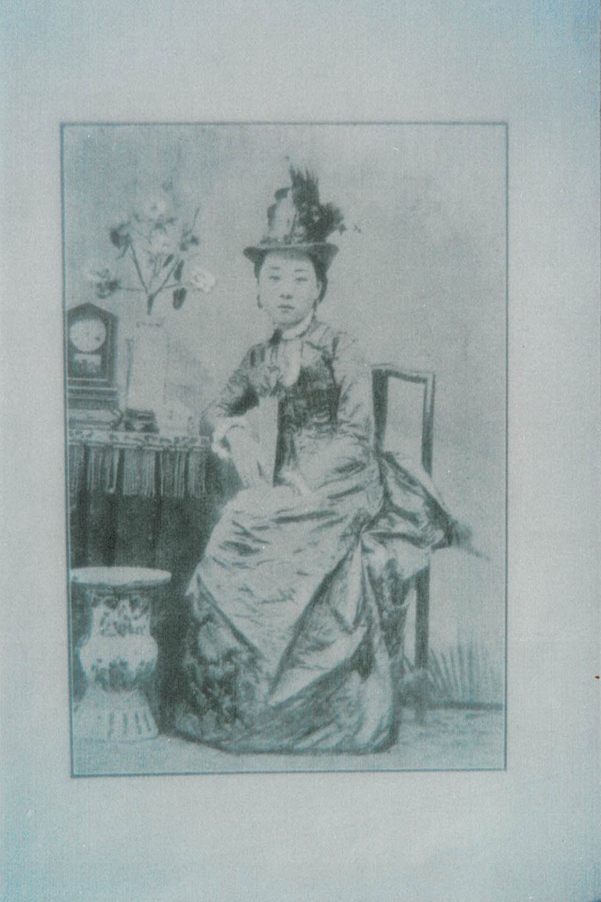 A black-and-white image shows an Asian woman wearing a formal nineteenth-century European gown and a feathered hat. She sits with one elbow resting on a table with a clock and tall vase of flowers, while a large decorative porcelain vase sits on the floor by her feet