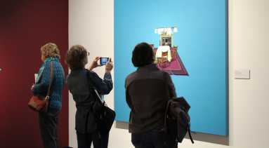 Three museum visitors observe works of art on the wall in a modern gallery. One wall is red and the other is white. One person takes a photo of a work of art with a cell phone.