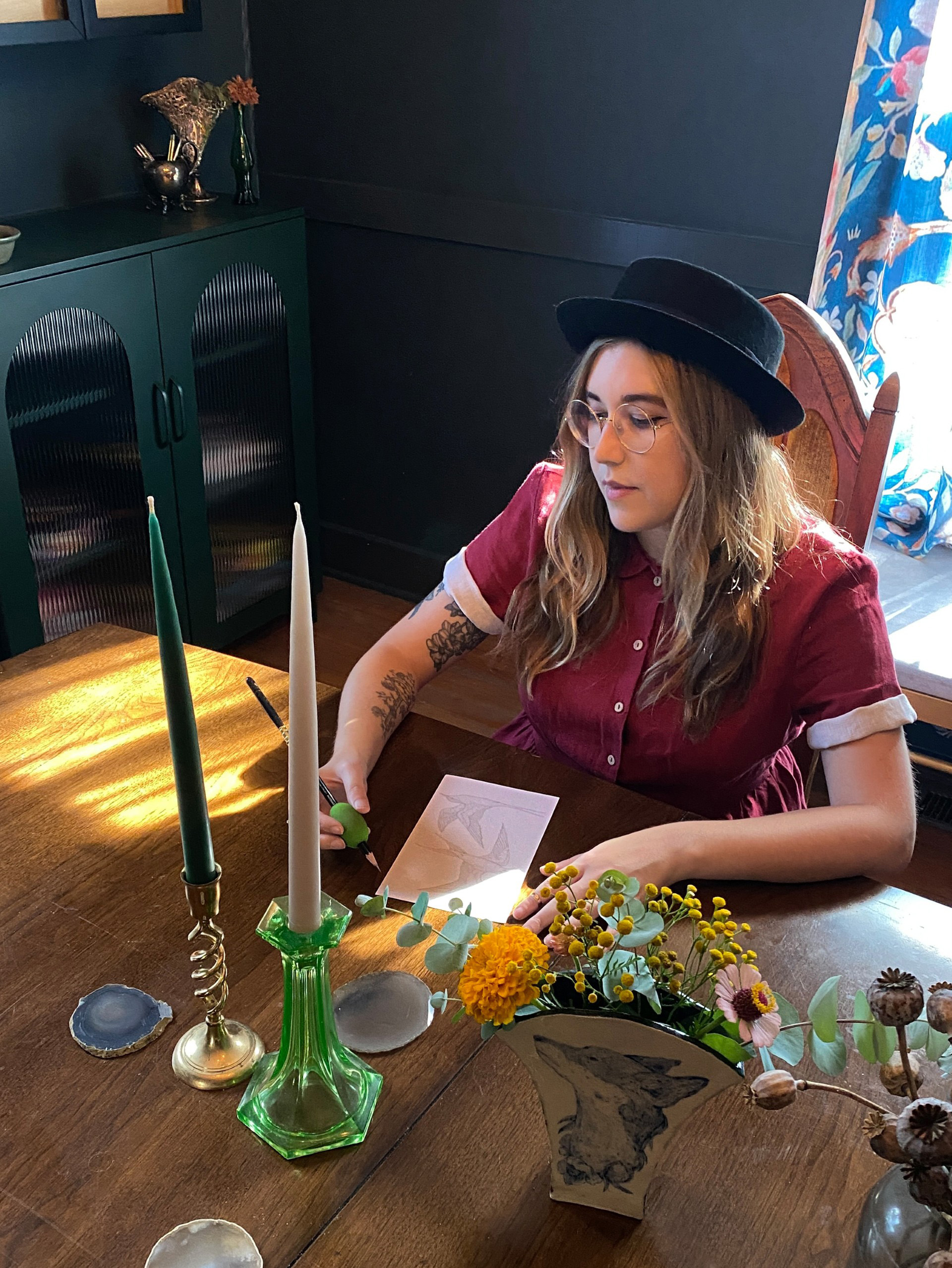 A young woman with light skin tone sits at a wood table and appears to be drawing on a small rectangular piece of paper. She wears a black hat and button-up shirt. The room's walls are painted dark.