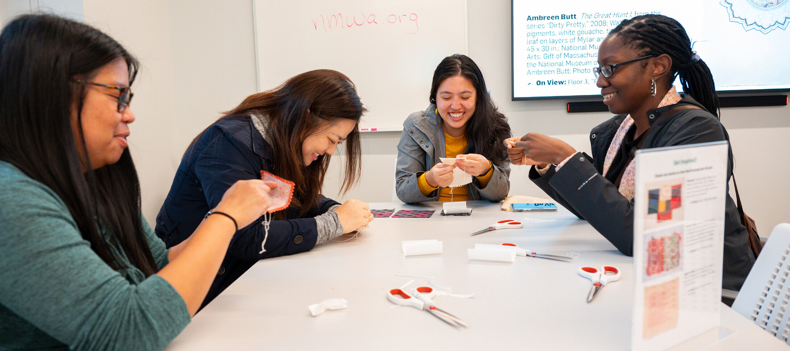 Four women sit at a table and hand-stitch white thread into white pieces of fabric.