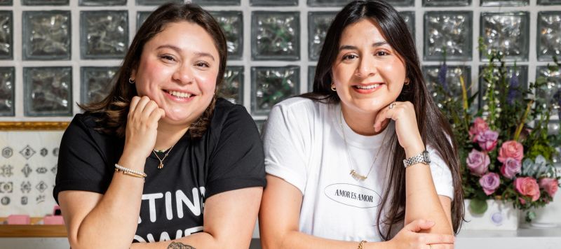 Two young women, both with straight dark brown hair, pose with their arms resting on a counter and one hand under their chin. They are under a fluorescent sign that says "JZD" in the design of a smiley face.