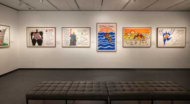 A contemporary art gallery with white walls displays colorful, framed artworks featuring handwritten text, patterns, and illustrations. A black cushioned bench sits in the room.