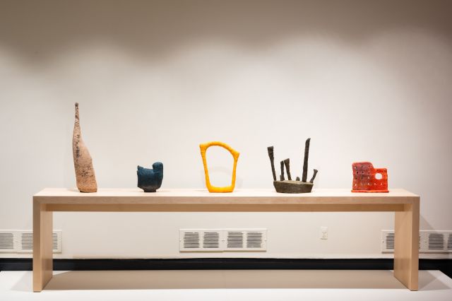 An installation view of five different colored abstract contemporary sculptures sitting in a line on a long light wood rectangular table that is on a low white platform.