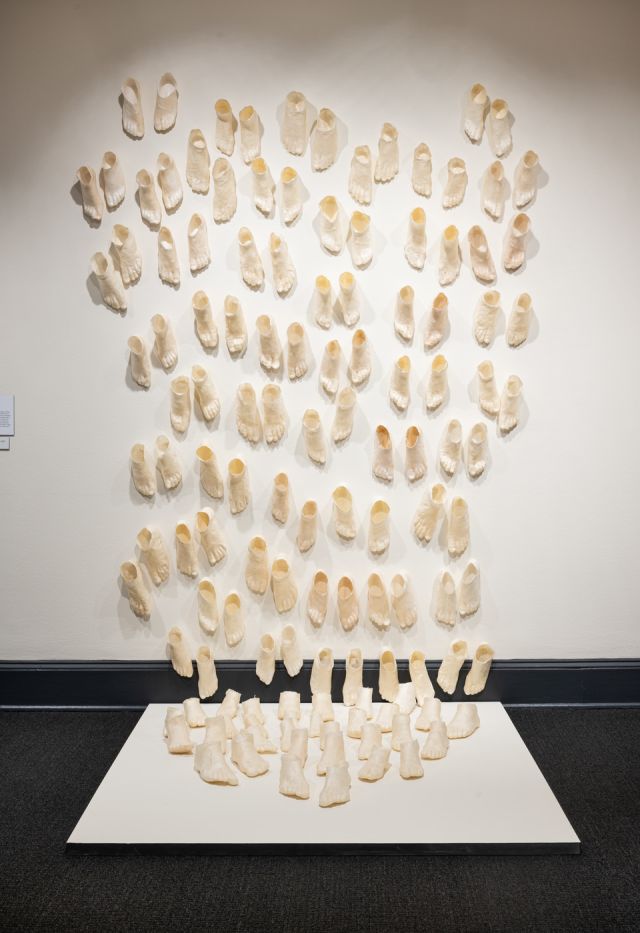 A contemporary art installation made up of countless beige foot sculptures that extend from the walls onto a low white platform on a dark floor. The feet are in pairs in varied sizes and staggered as if moving.