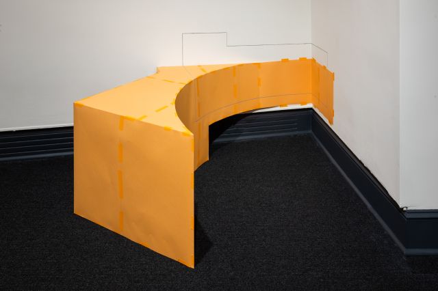 An orange geometric abstract sculpture with varied angles positioned against a corner on the floor. The sculpture has painted lines extending onto the walls.