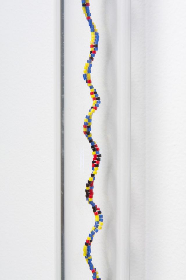 Detail image of a sculpture of a glass tube affixed to a white wall with a spiraling column of small, multicolored glass beads. The beads are blue, red, yellow, green and black.