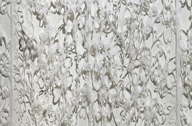 Detail view of delicate, white tissue paper that has been cut with intricate floral patterns which create shadows on the wall behind it.