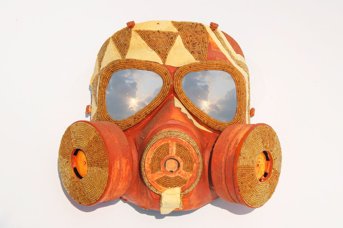 A gas mask decorated with shiny seed beads, animal hide, paint, and Oklahoma red dirt. The added materials form a geometric pattern on the forehead area, and a clouded sky can be seen reflected in the eye lenses of the mask.