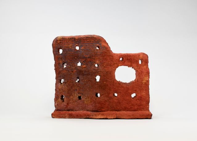 An irregular shaped red abstract sculpture that looks like the side wall of an old building structure. Rows of holes and one bigger void space cover the surface of the grainy, rough sculpture.