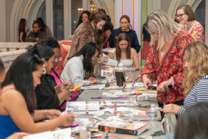 A group of women gathered around a long table work on arts and crafts projects, using magazines, scissors, and glue sticks in a bright, ornate indoor setting.