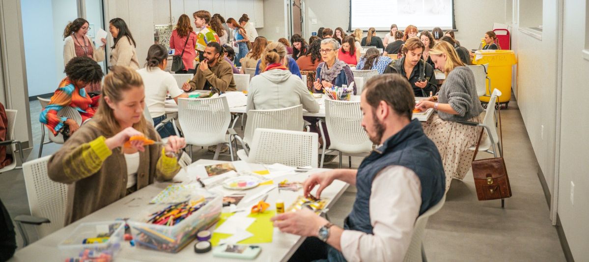 A large group of people sit at tables in a bright, modern classroom, working on arts and crafts projects with colorful supplies. Others stand and talk in the background. A presentation is displayed on a screen.