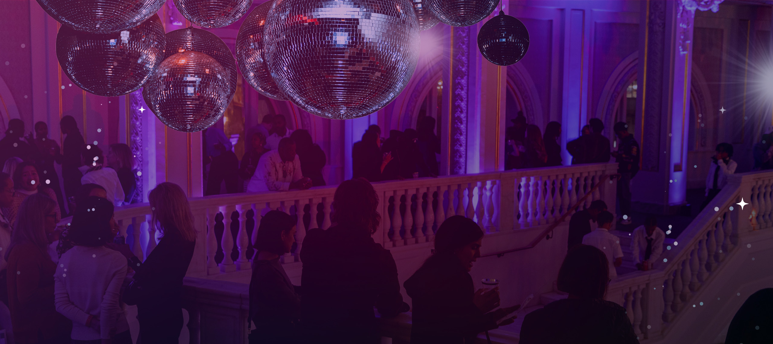 A crowd of people socializes along a grand staircase and balcony inside an ornate building, illuminated by purple and pink lighting. A cluster of disco balls hangs from the ceiling.