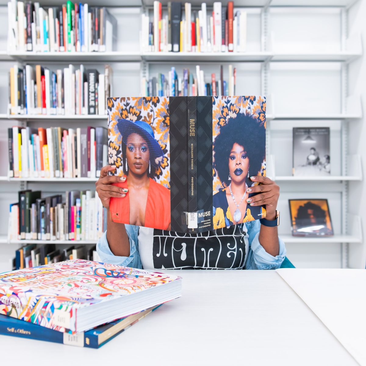 A dark-skinned person sits at a white table in front of shelves of books. They hold up a large art book, which obscures their face, and features the portraits of two elegant and stylish Black women on the cover and back.