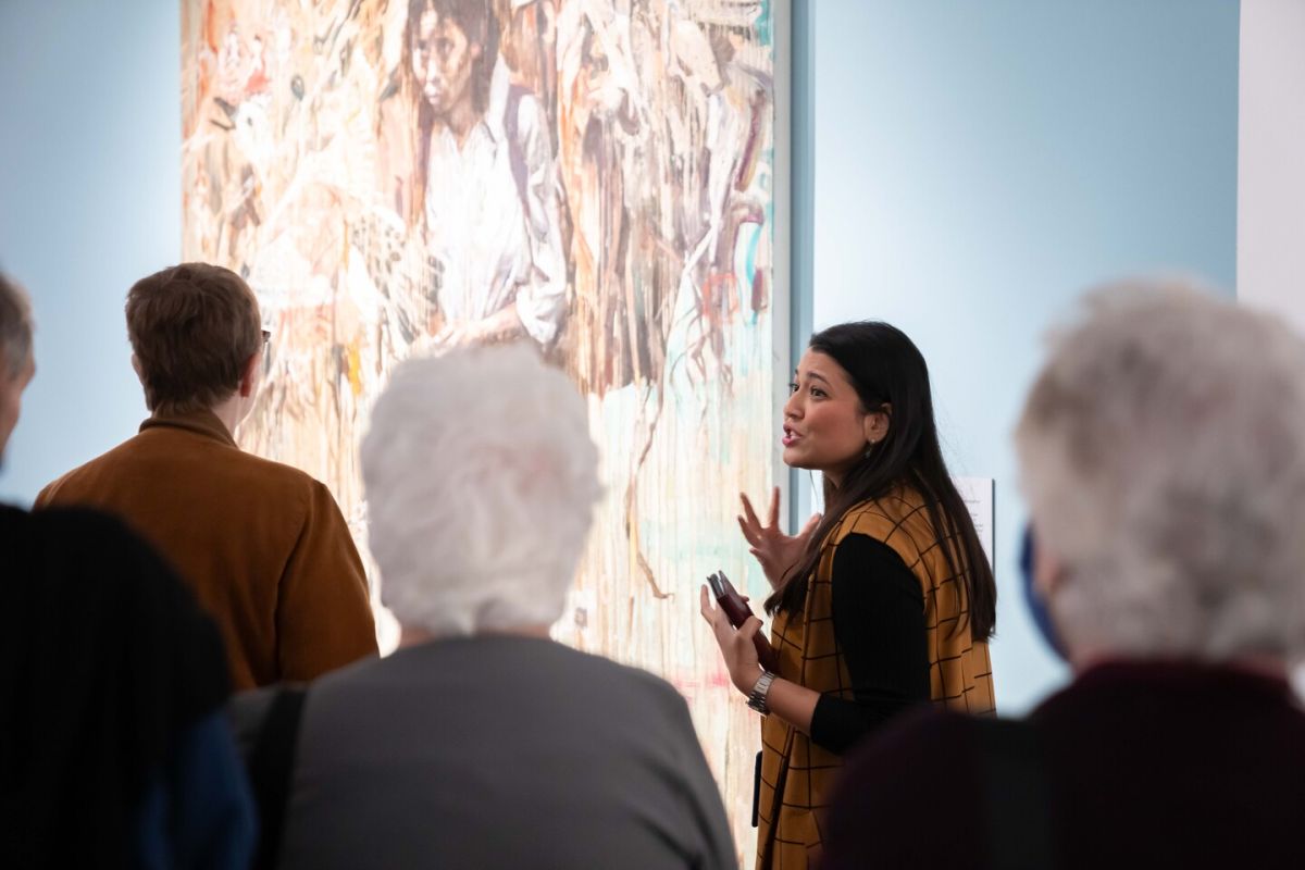 A woman is standing in front of a group next to a large painting. She is gesturing with her hands as she talks, engaging the group. She has a medium-light skin tone, long hair, and is wearing a mustard yellow dress.