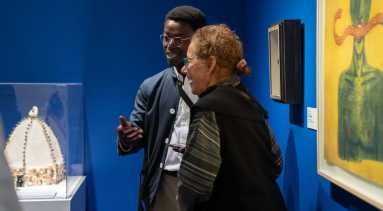 Two museum visitors in conversation next to an art piece. A man with a dark skin tone, glasses with a golden frame wearing a navy blazer is talking to a woman with a medium-light skin tone wearing a black jacket.
