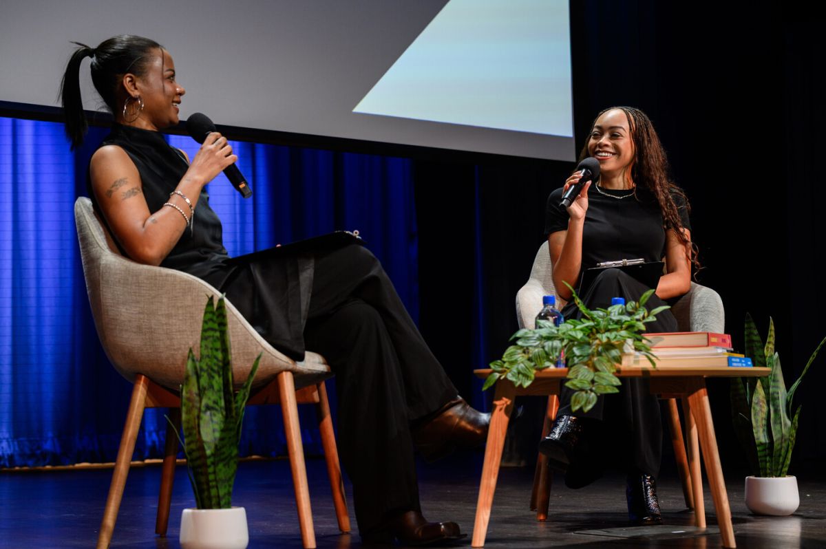 Two women sit in chairs on a stage, speaking into microphones. Both are wearing black outfits. There are plants and a stack of books on the table between them. Blue curtains and a screen are in the background.