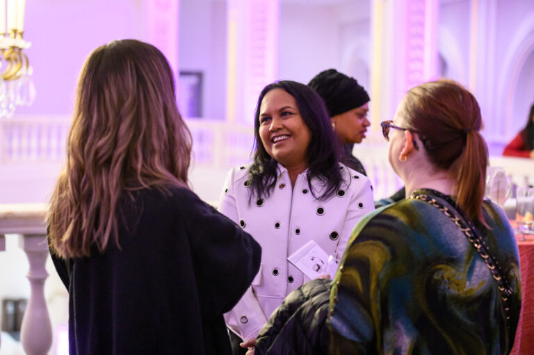 Three women engaged in conversation at an indoor event. The central woman smiles, wearing a white coat with black buttons. The others have long hair and glasses, wearing dark and patterned clothing. The background is softly lit with purple hues.