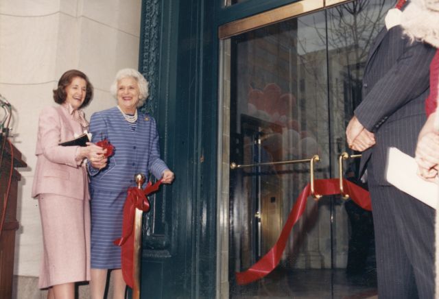 Two older light-skinned women in suit and skirt sets smile and cut a red ribbon on the doors of an institution. One woman has brown hair and the other hair white hair.