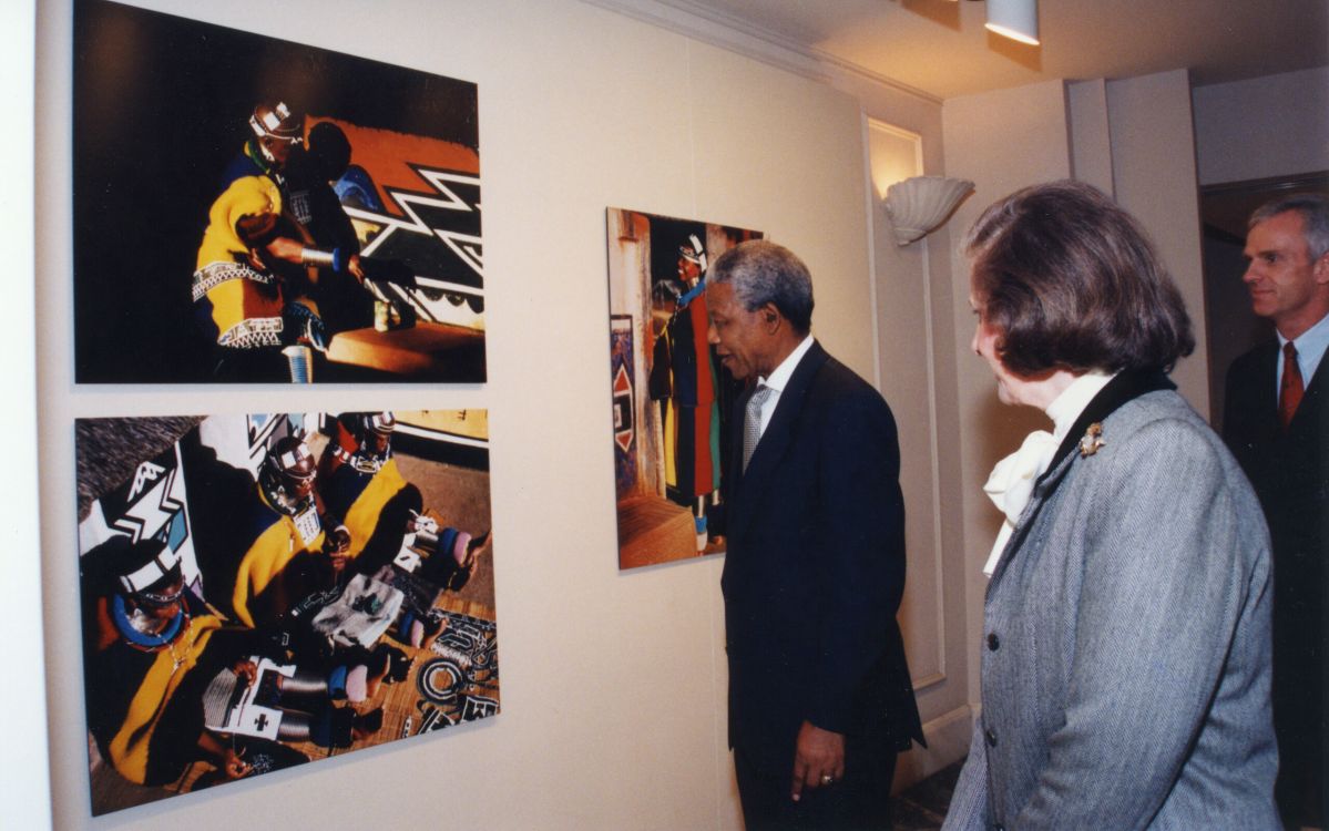 Three people in formal attire view colorful, abstract artwork displayed on a gallery wall. Two art pieces show people in vibrant, patterned clothing. The group stands attentively, observing the art.