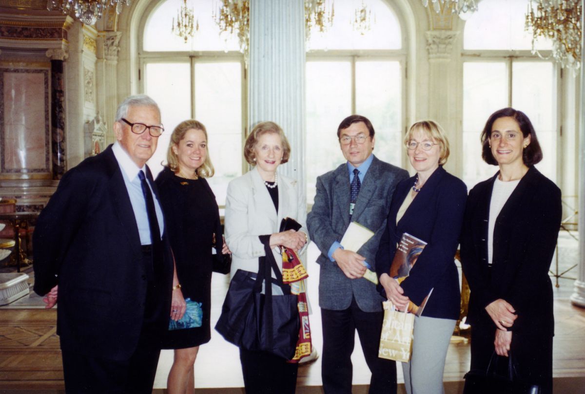 A color photograph of a group of 2 light-skinned men and 3 light-skinned women who stand in an ornate room, with large windows, crystal chandeliers, colored marble details, and gold accents. The figures are all wearing business attire and smiling at the camera.