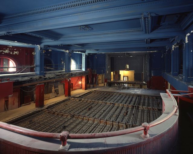 A large, ornate hall is photographed from a balcony, looking down onto a floor under renovation, with steel supports in place. Around the hall paint chips off of the walls and a layer of dust coats the balcony railing. Construction debris is visible on the left side.