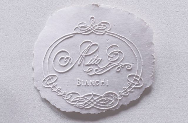 The name ‘Rita Bianchi’ is inscribed in a cursive typeface and slightly raised off the surface of a thin white paper pulp base with frayed edges.