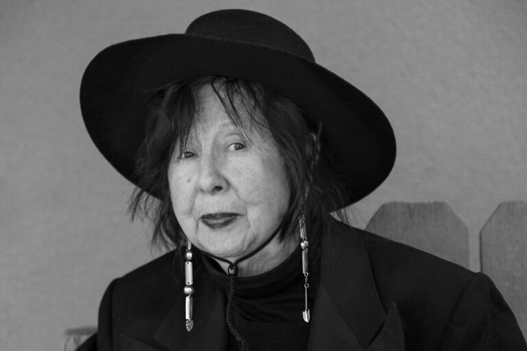 A black and white photograph of a light-skinned Native American woman with short dark hair and dark lipstick. She is dressed in all black and is wearing a black wide-brimmed hat and long silver earrings made of rectangular metal shapes.