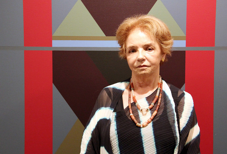 An older woman with light skin tone and short, light brown hair stands in front of an abstract painting. She wears a black and white striped shirt with two beaded necklaces. The painting behind her depicts grey and red rectangles framing grey, brown, and green triangles.