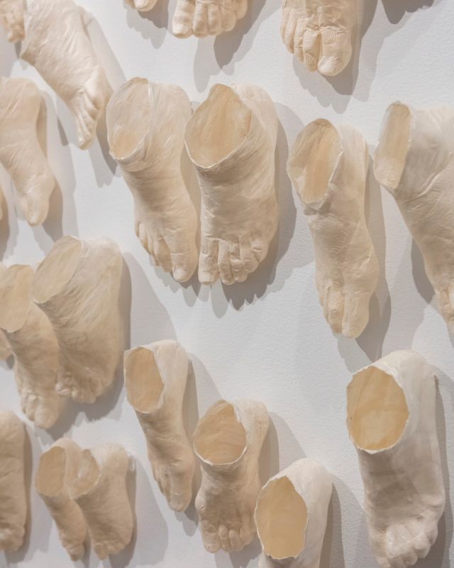 Detail view of a contemporary art installation made up of countless beige foot sculptures that extend from the walls onto the dark floor. The feet are in pairs in varied sizes and staggered as if moving. Marks on the sculptures indicate the folds of human skin and bone.