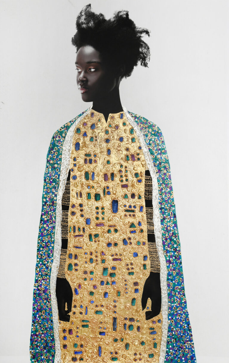 A photograph depicting a woman with dark skin and natural hair standing against a light background, wearing a cape and robe adorned with intricate gold patterns and colorful gemstones.