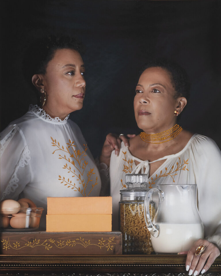 A photograph of two women in embroidered white tops stand behind a table with eggs, orange boxes, pasta in a jar, and a glass pitcher of milk, set against a dark background.