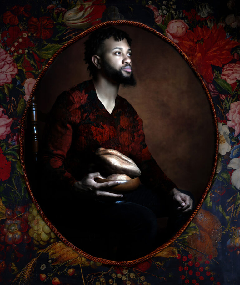 A photograph of a Black man with a beard wearing a red and black shirt, holding a bowl filled with sweet potatoes, and looking towards the right. The background of the photograph is a painted brown color. The photograph is surrounded with braided red trim and a floral background.