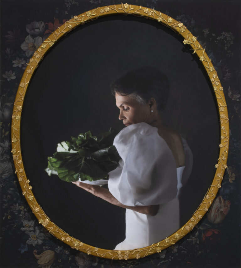 A photograph of an older Black woman with short grey hair in profile. She wears a long, white, gown with puffy sleeves. She holds and gazes at a head of collard greens.