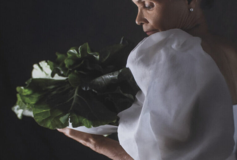 A photograph of an older Black woman with short grey hair in profile. She wears a long, white, gown with puffy sleeves. She holds and gazes at a head of collard greens.