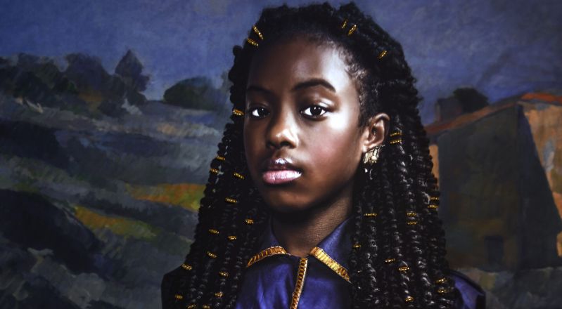 A photograph of a young Black girl. She is seated and wearing a blue dress with gold accents. She holds in her lap a doll, which is dressed in a gold dress and headwrap. The background is a landscape painting by Cezanne.