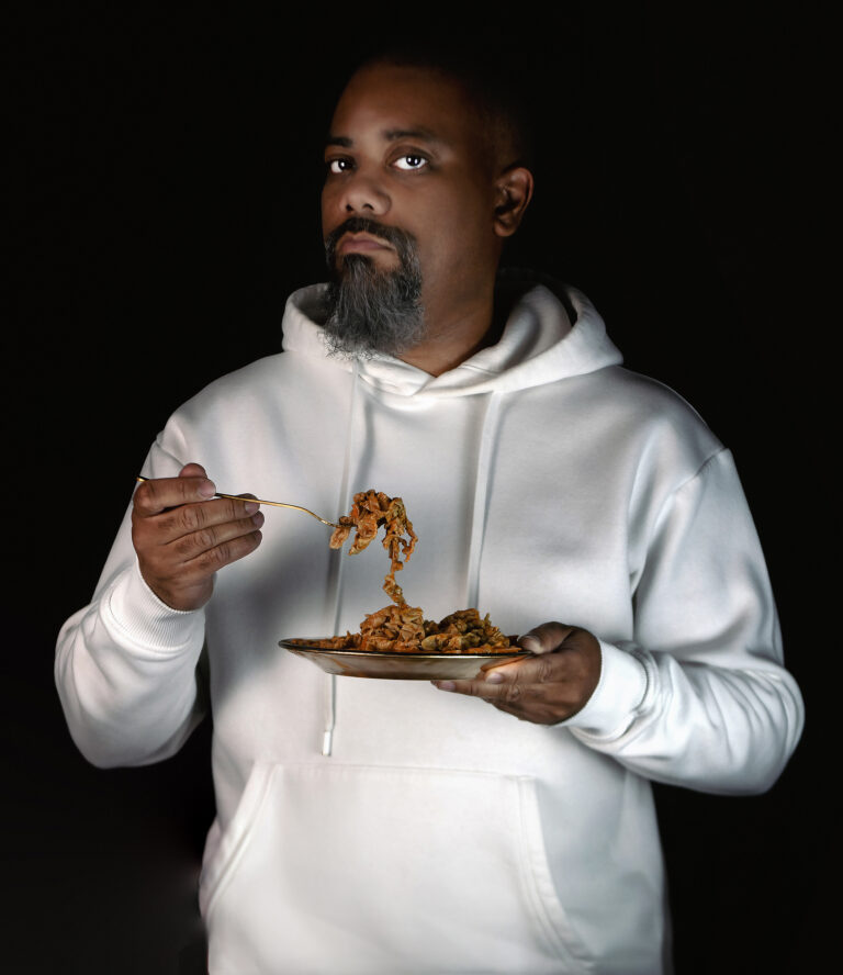 A photograph shows a Black man with facial hair wearing a white hooded sweatshirt. He holds a plate of chitlins in one hand. In the other hand he holds a fork loaded with chitlins.