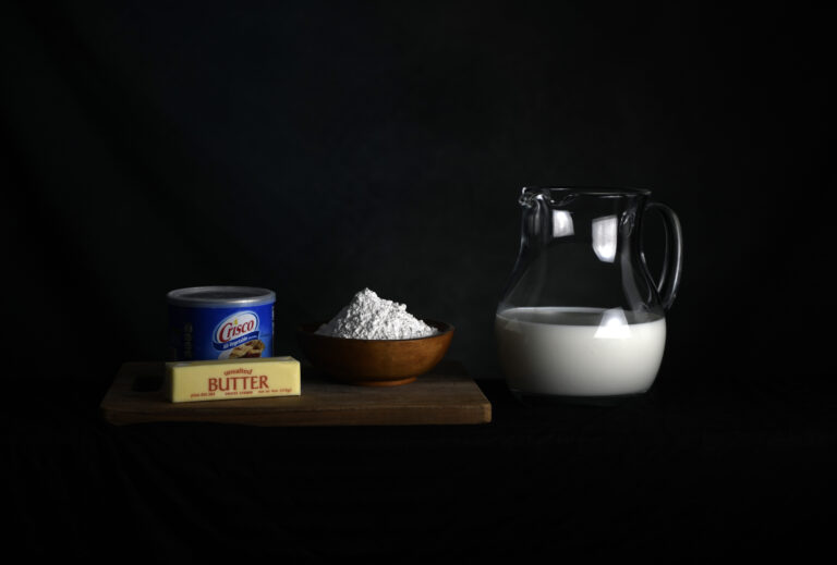 A photograph shows a tin of Crisco, a stick of unsalted butter, and a bowl of flour atop a wooden cutting board. A glass pitcher of milk rests next to the other ingredients.