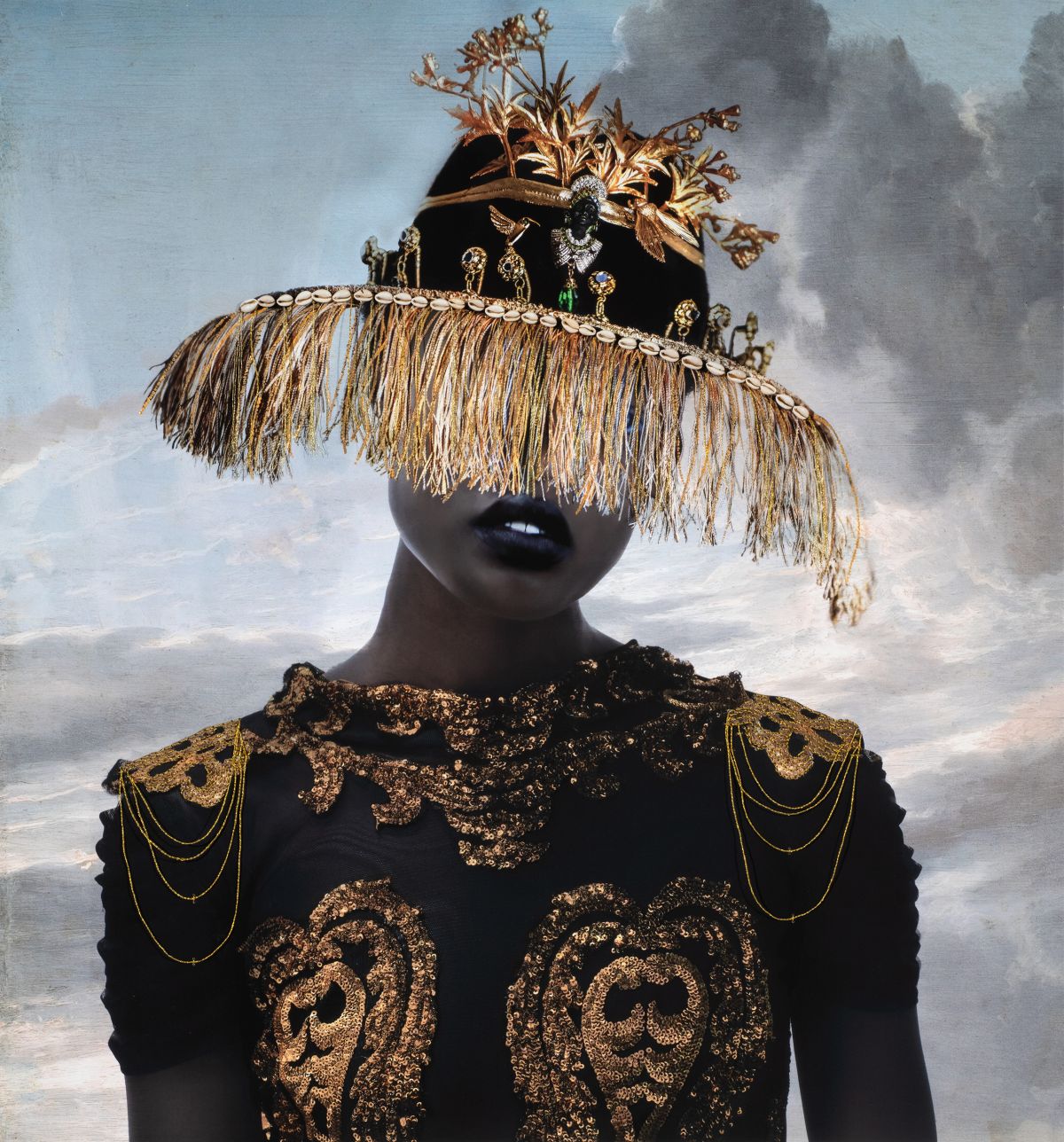 A person with dark skin tone in a black-and-gold ornate dress with gold chain details wears a wide-brimmed hat decorated with gold fringe and plants, obscuring their eyes. The background shows a cloudy sky.