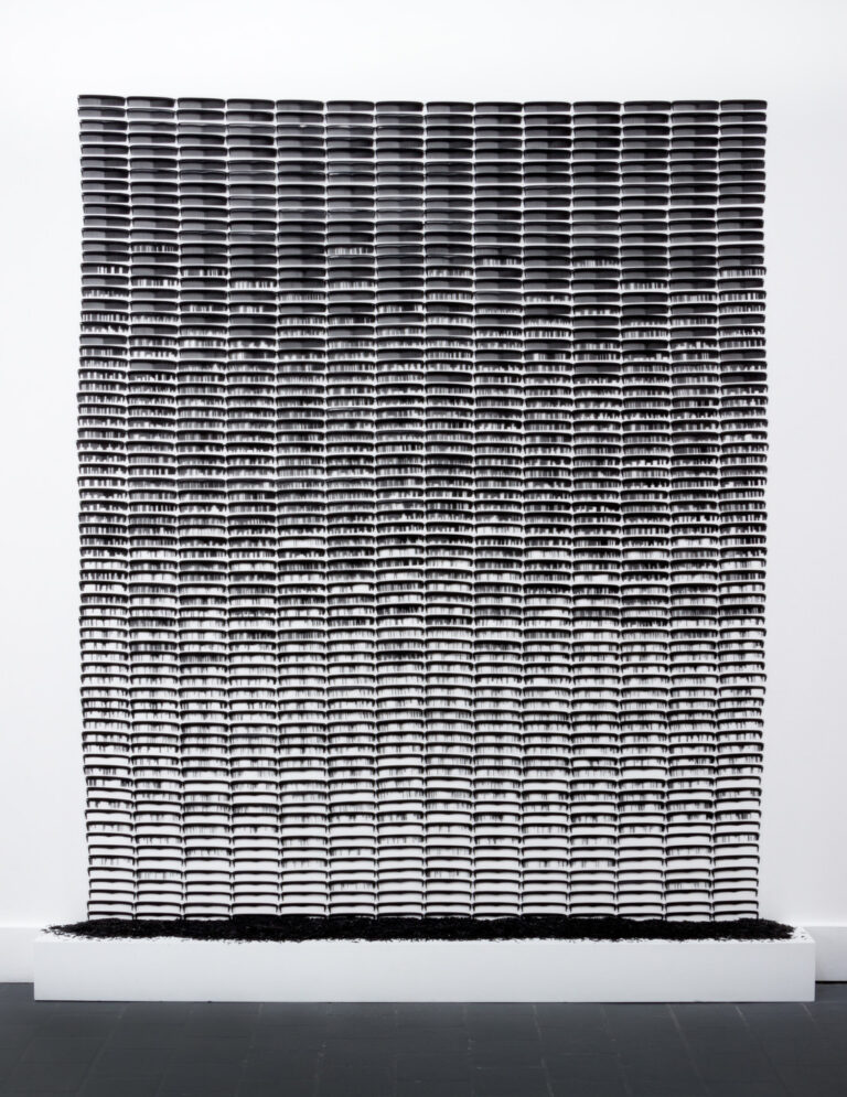 A large rectangular sculpture made of black, plastic pocket comes attached side by side in rows against a white background. As the combs descend, more and more of their plastic teeth are removed, leaving gaps and open spaces. The removed pieces of comb teeth are pooled at the base of the sculpture.