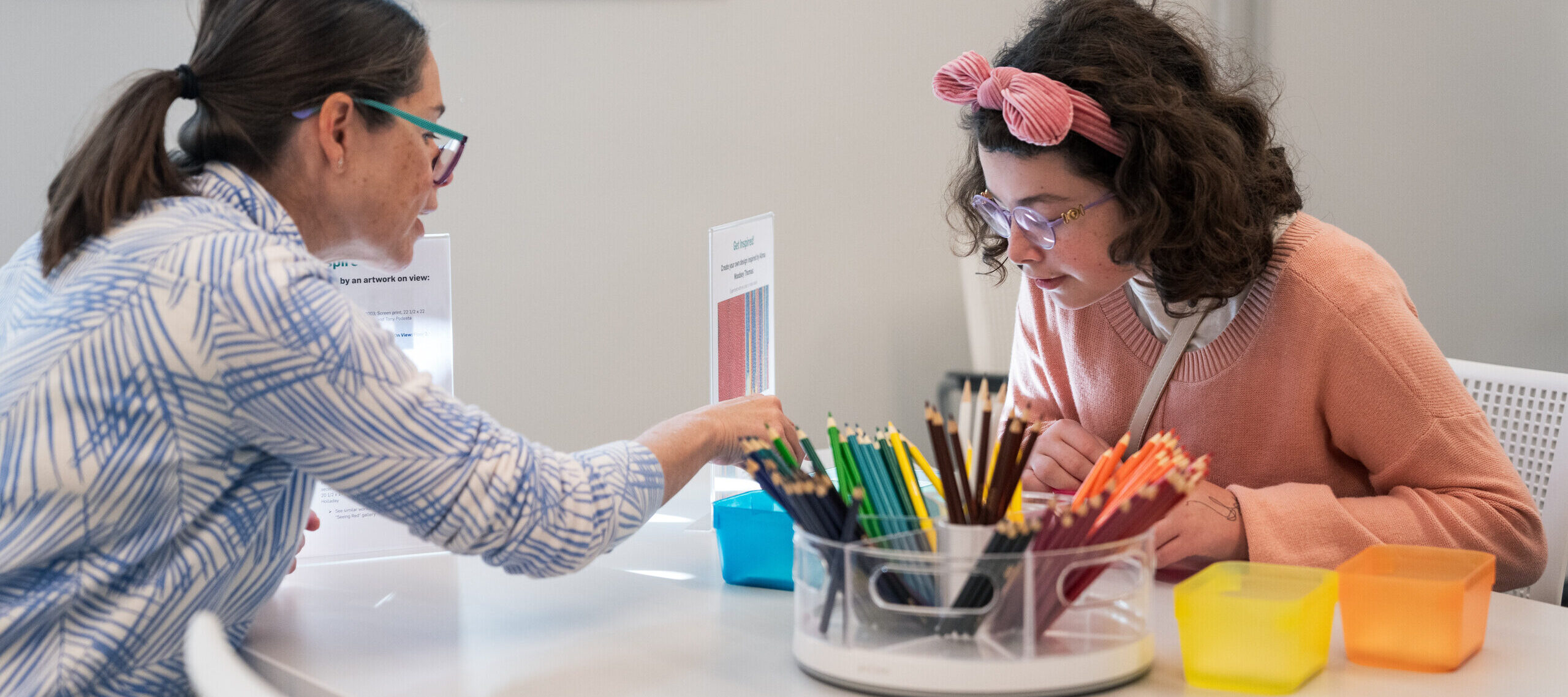 Two light-skinned women sit at a table and color with colored pencils.