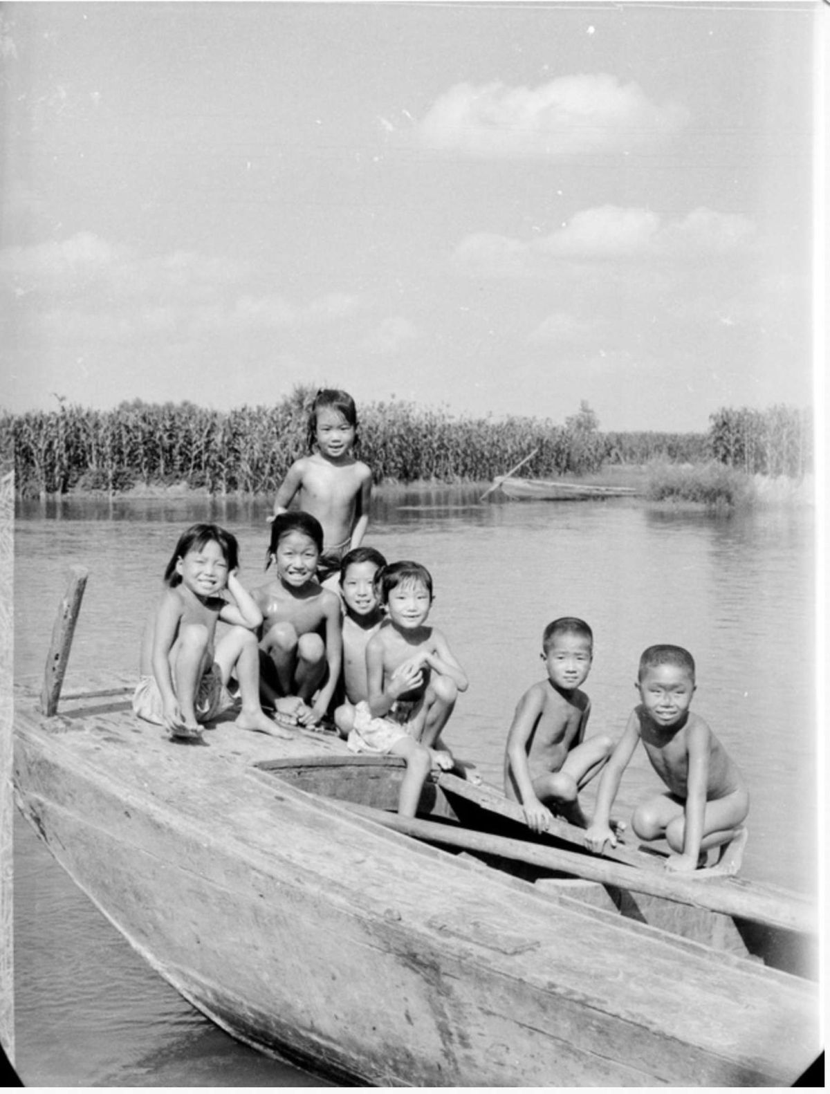 A black-and-white image shows a group of nude and half-clothed Asian children on a boat on a river. They stand and crouch in a group to pose for a picture, smiling at the camera.