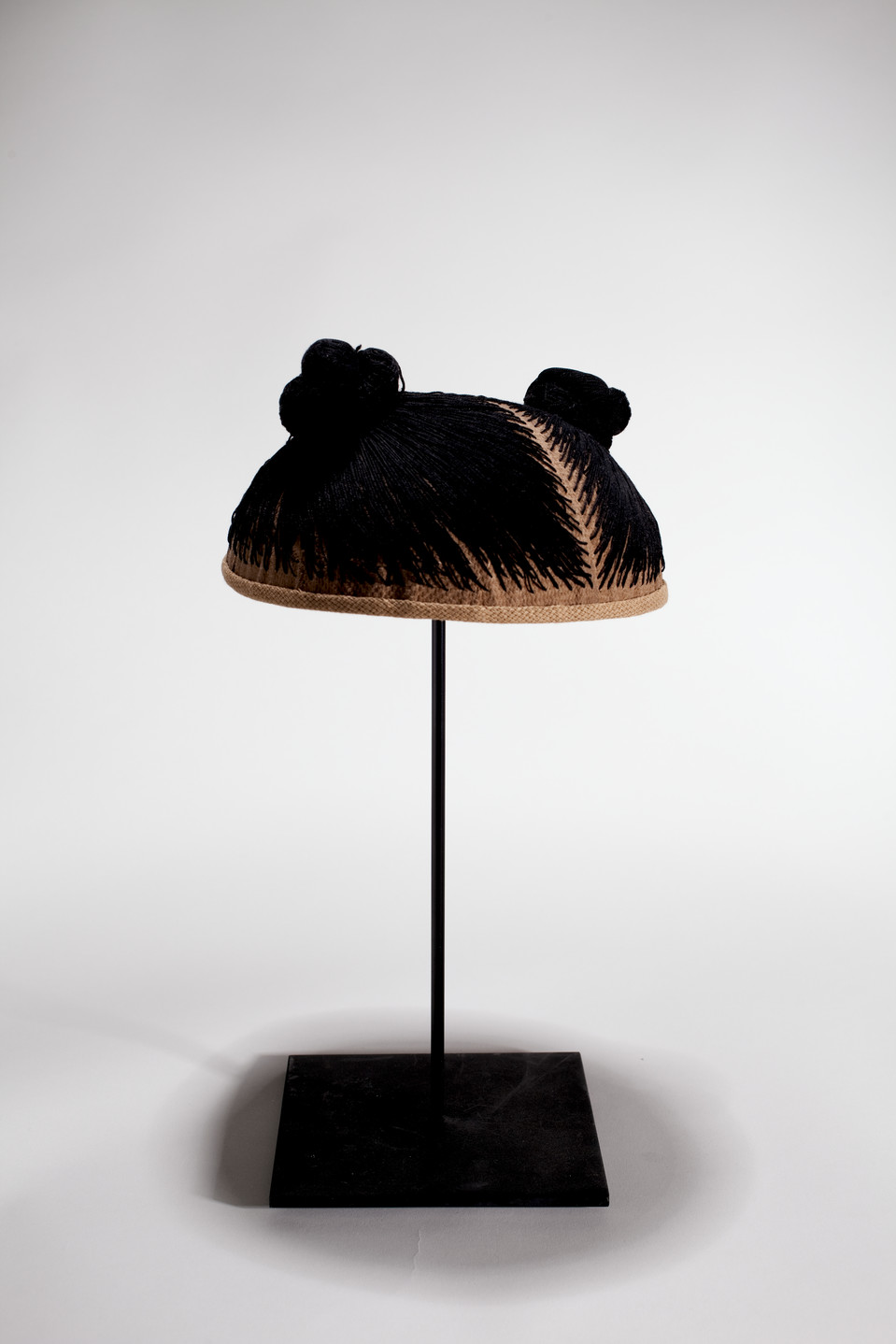 A wig cap sits mounted on a black stand. Black thread is stitched into the cap, mimicking hair. The thread forms two small buns.