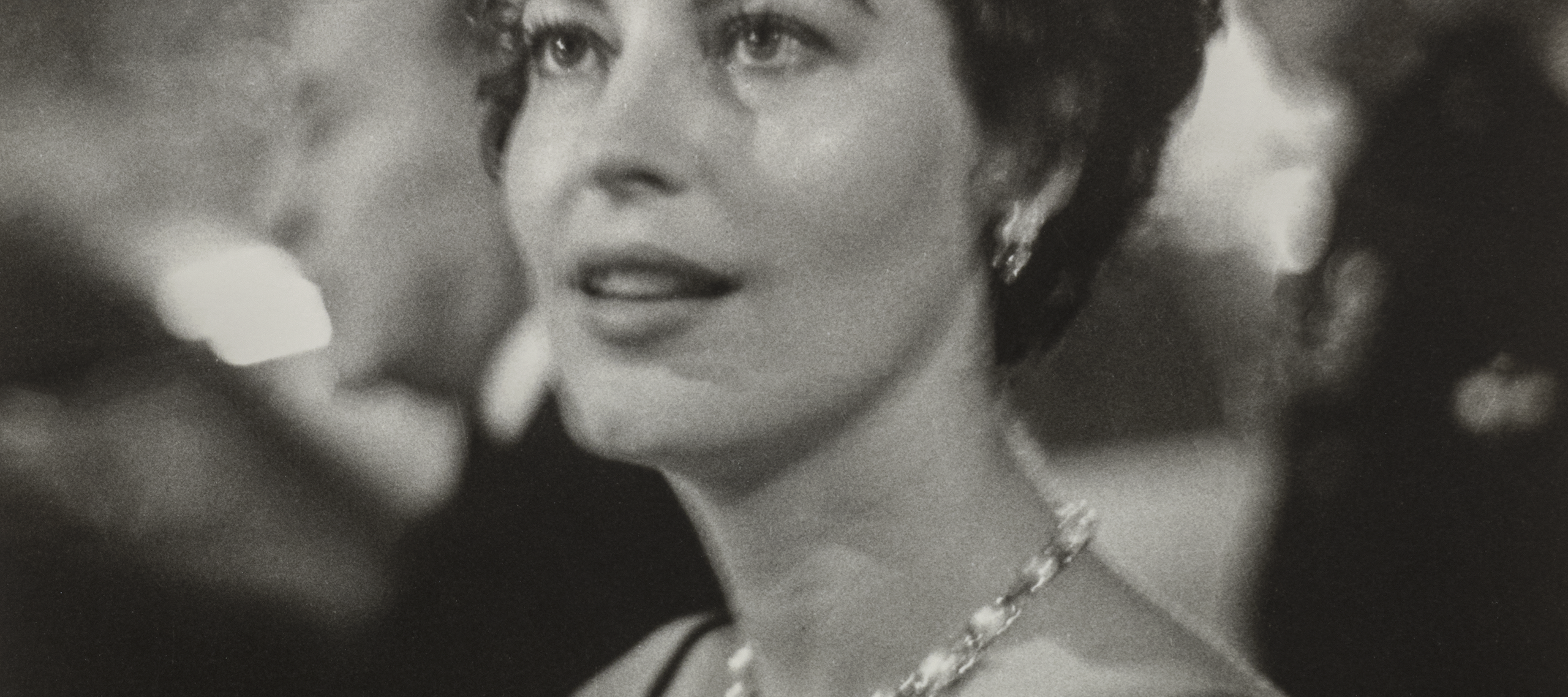 Ava Gardner, a woman with light skin tone and wavy, short brown hair, is shown from the shoulders up. She wears a jeweled necklace and is surrounded by lights and people at a party. She looks to the side of the camera at something out of frame with a fixed gaze and slight smile.